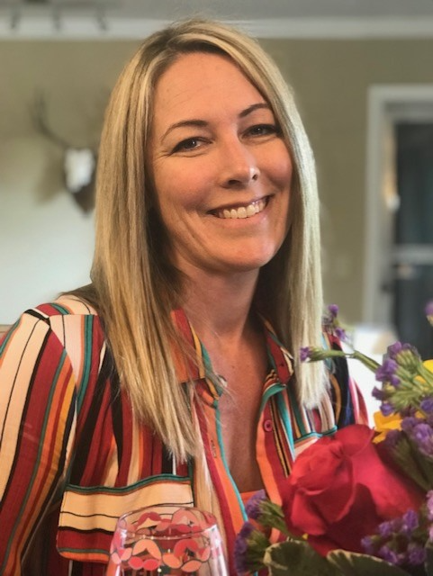 A white woman with straight blonde hair has a giant smile on her face and is looking into the camera, she is wearing a colorful striped collared shirt.